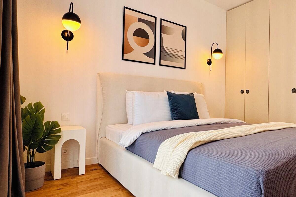 A bedroom with a bed, bedside lamps, a small white nightstand, a potted plant, and a wardrobe.