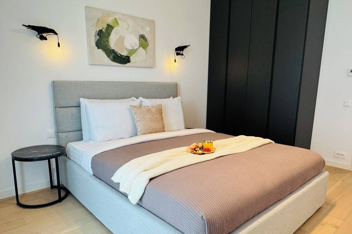 A modern bedroom with a bed, bedside tables, a painting, and wall-mounted lights.