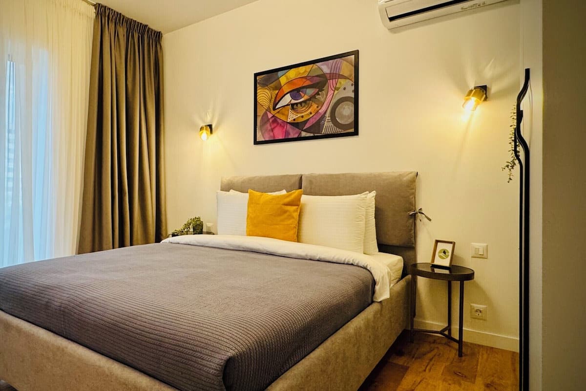 A bedroom with a bed, curtains, a painting, wall sconces, and an air conditioning unit.