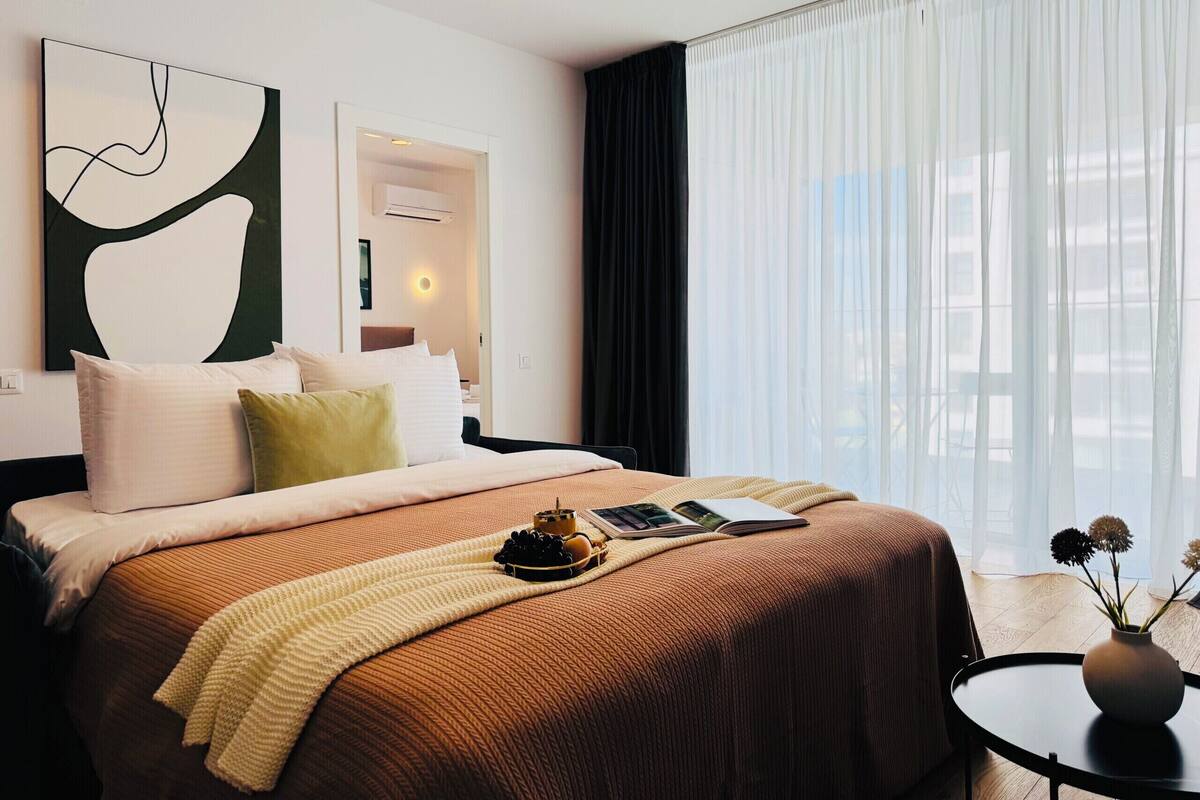 A hotel room with a bed, a bedside table, a window with sheer curtains, and a wall-mounted artwork.