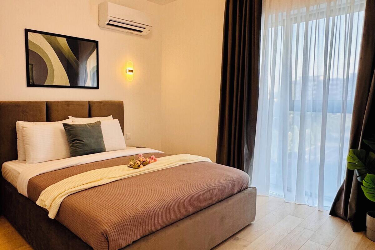 A hotel room with a bed, bedside tables, an air conditioning unit, a painting, and a window with curtains.