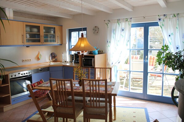 Une cuisine avec des meubles en bois, une table à manger avec des chaises, une lampe suspendue et une vue à travers une porte vitrée.