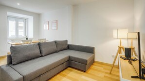 A modern living room with a grey sectional sofa, a wooden dining table with chairs, a floor lamp, and a flat-screen TV on a stand. - Nigrum Apartment, Santos, Lisbon (Lisboa)