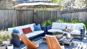 Un patio con un sofá blanco, dos sillas de madera y una mesa baja con un jarro de metal.
