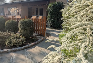 Un chemin pavé mène à un bâtiment entouré d’une clôture en bois, et l’ensemble est agrémenté d’arbustes bien entretenus et de plantes en fleurs.