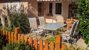 Een terras met een tafel en stoelen, een houten omheining en een huis met een gestreepte luifel.