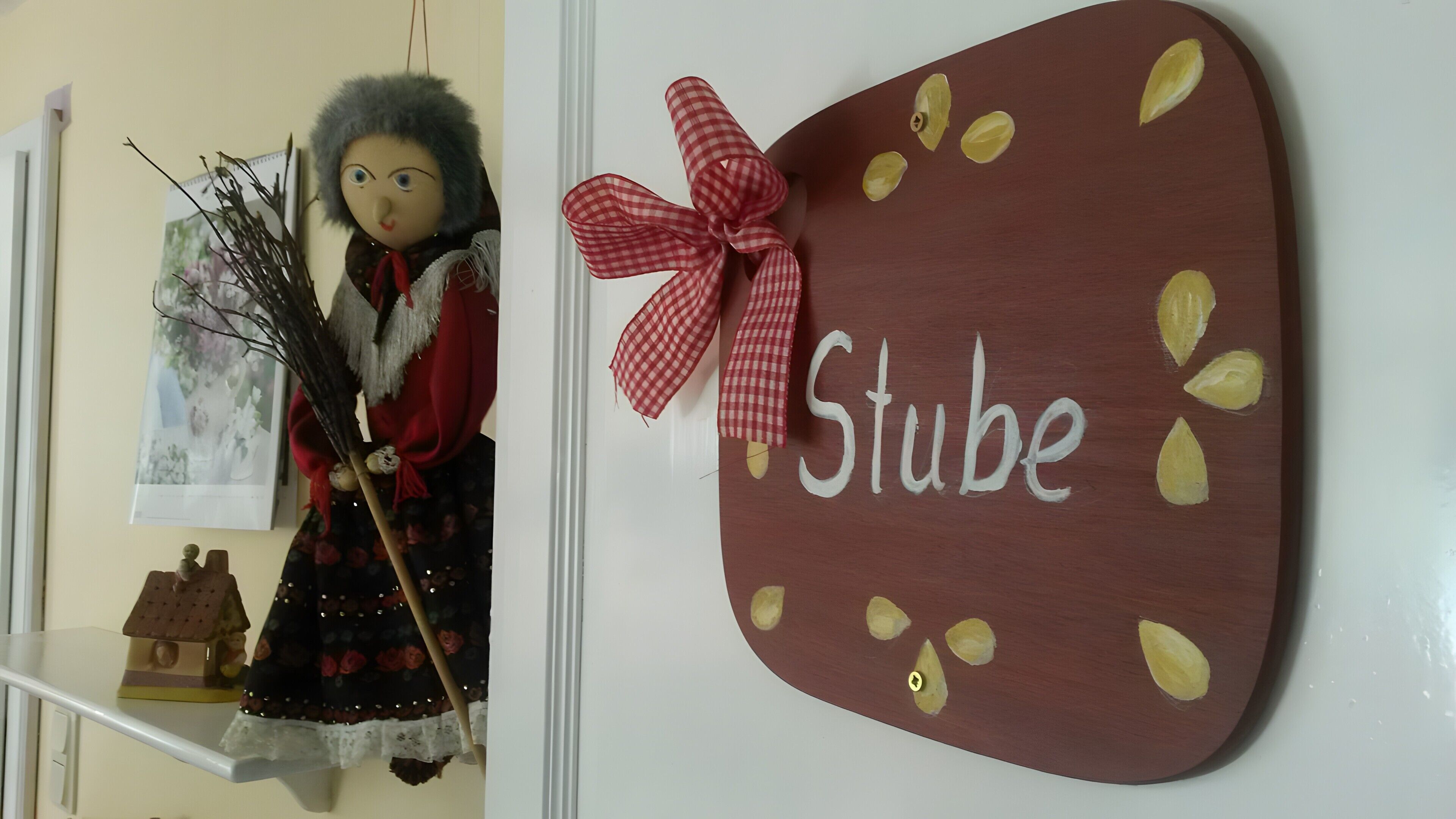 A doll with a red scarf and a broom, a wooden sign with 'Stube' and leaves, and a small house model on a shelf.