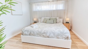 A bedroom with a bed, two bedside tables, a window with sheer curtains, and a wall-mounted picture. - JB’s - Jetty Busselton Stay (West Busselton)
