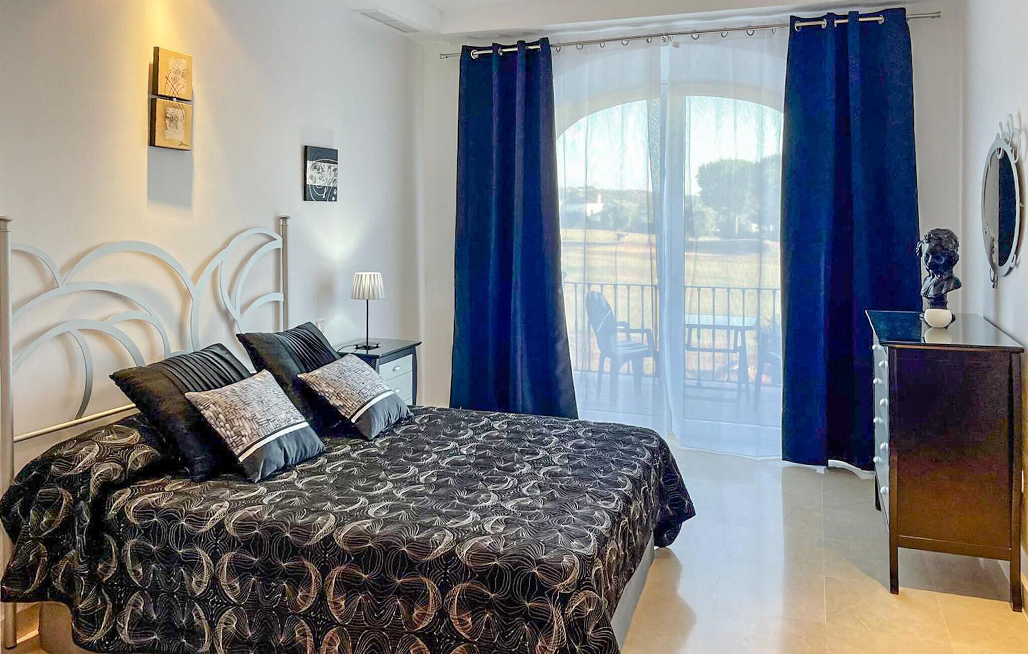 A bedroom with a bed, a nightstand, a dresser, a mirror, and a view of the outdoors through a large window with blue curtains.
