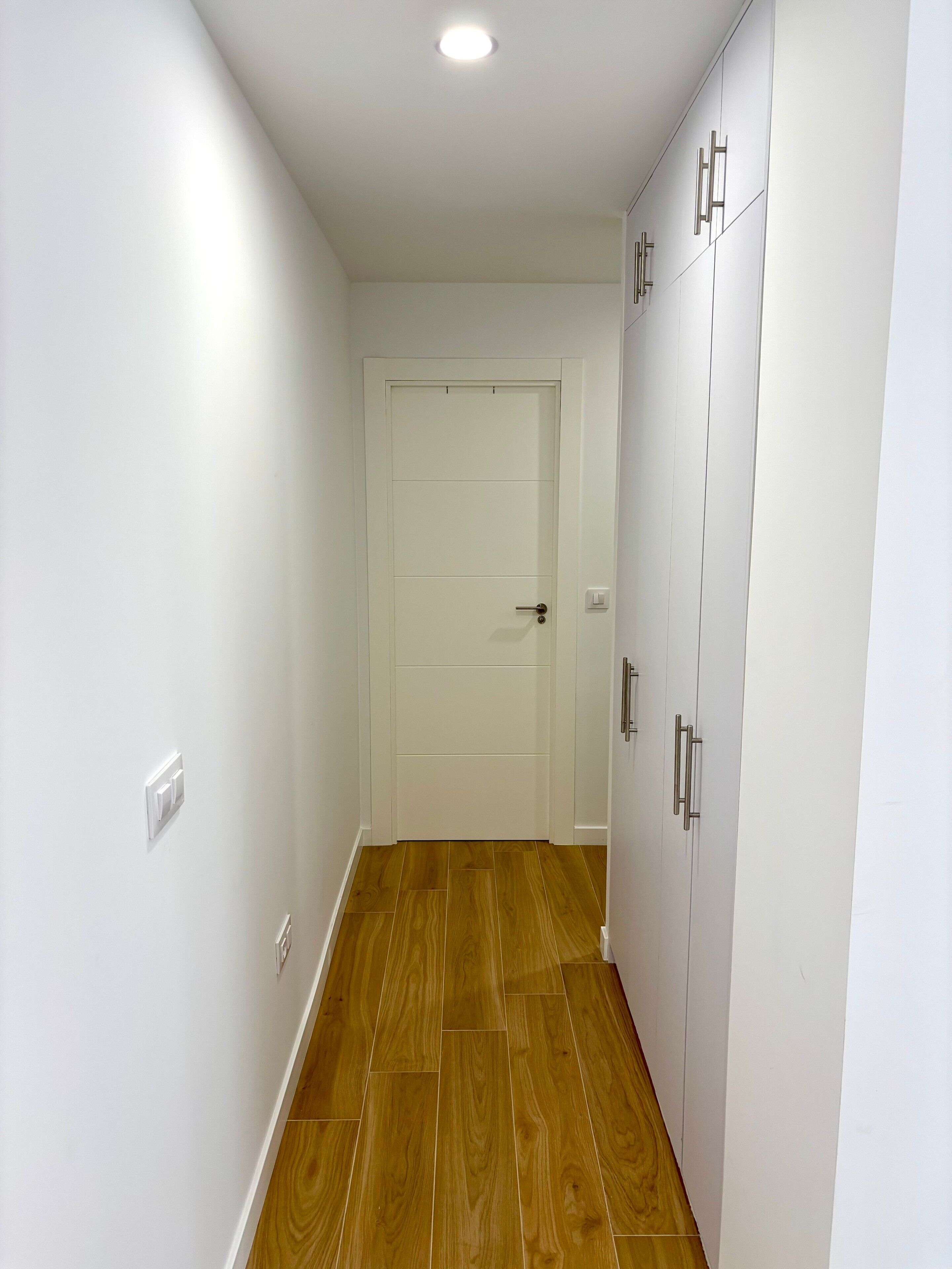 A hallway with a wooden floor, white walls, a white door, and a wardrobe with silver handles.