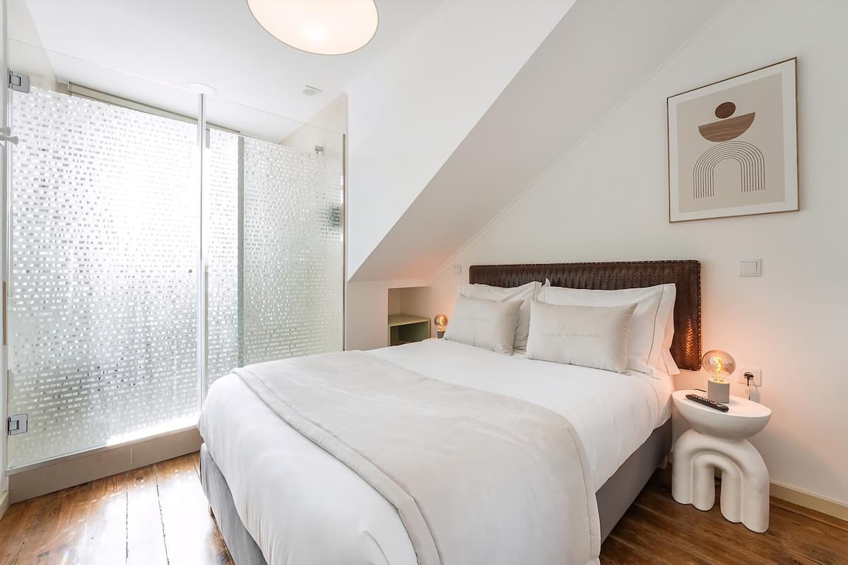 A bedroom with a bed, bedside tables, a nightstand, a wall-mounted light, and a glass door leading to a shower area.