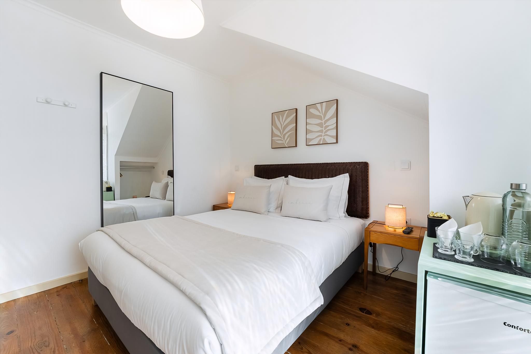 Habitación de hotel ordenada con una cama grande, lámparas de noche, un espejo y una mesita con vasos y una jarra.