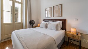 Un lit bien fait, avec du linge de lit blanc, une table de chevet avec une lampe et un ventilateur fixé au mur.
