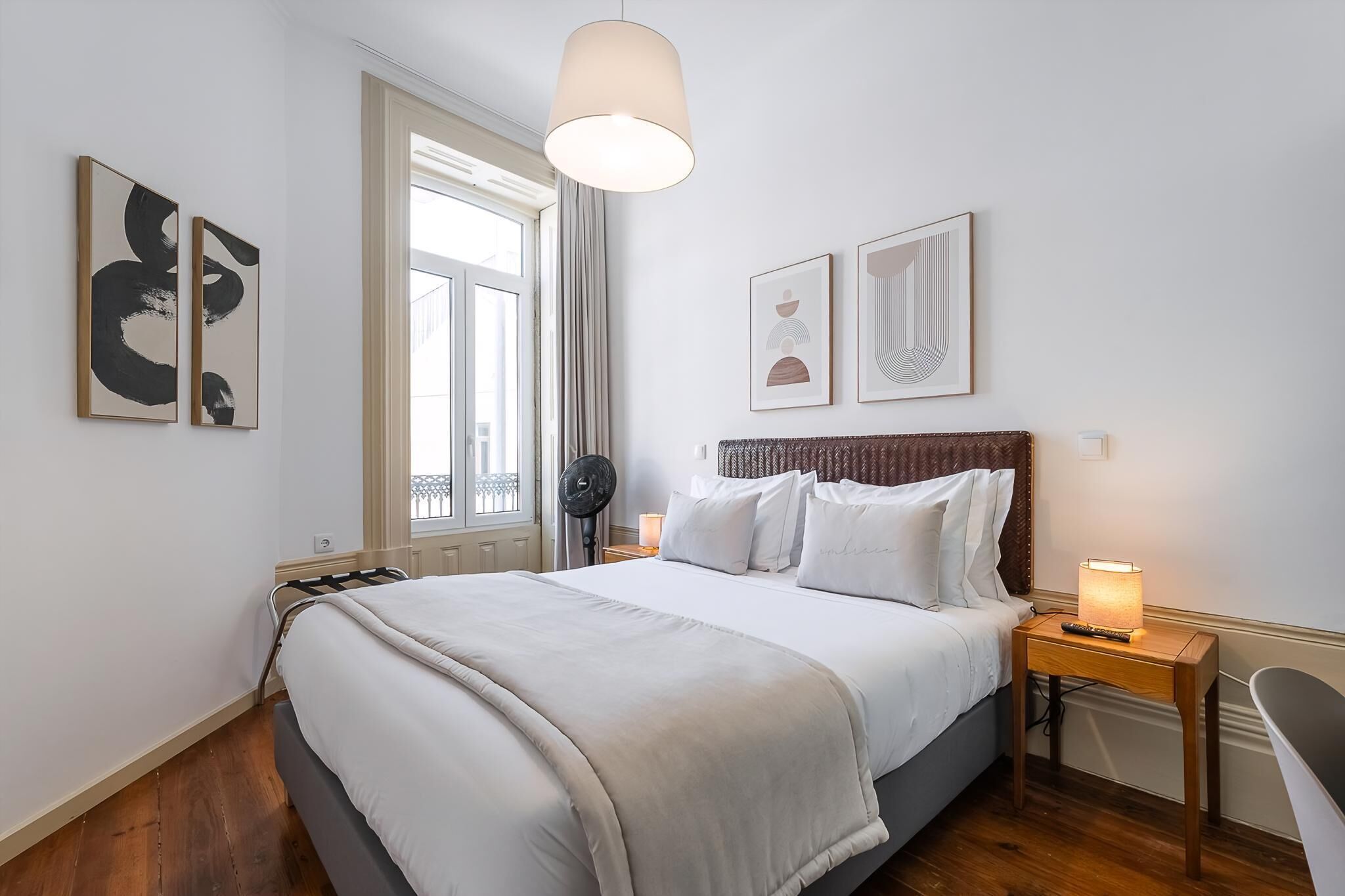 A bedroom with a bed, a bedside table with a lamp, a fan, and artwork on the walls.