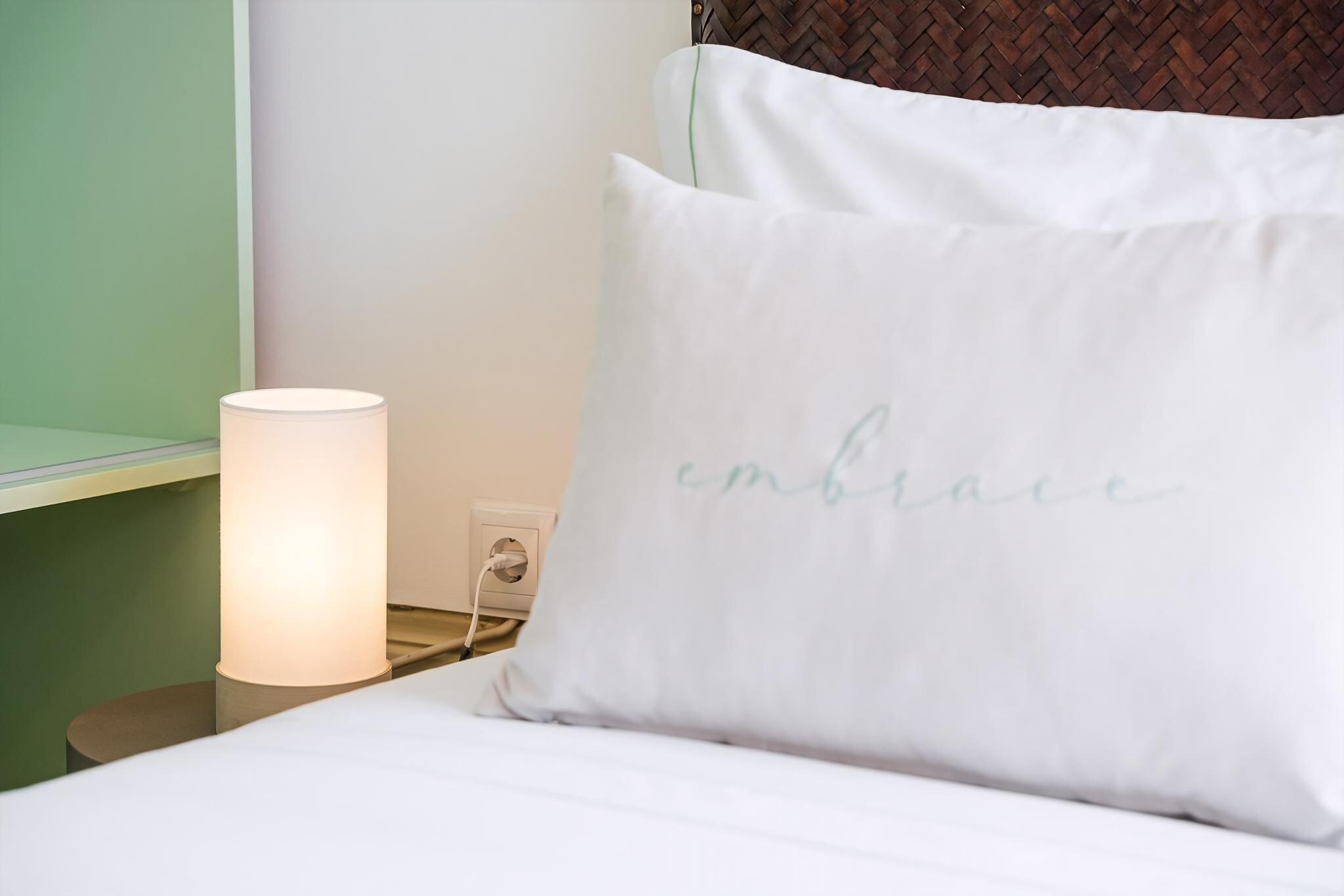 A bed with a white pillow with 'embrace' written on it, a bedside lamp, and a power outlet on a nightstand.