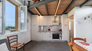 Ett kompakt kök med vita skåp, en mikrovågsugn och en diskmaskin. Här finns ett matbord med stolar, ett fönster med utsikt över byggnader och ett tak med synliga takbjälkar.