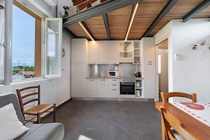 A compact kitchen with white cabinets, a microwave, and a dishwasher. There is a dining table with chairs, a window with a view of buildings, and a ceiling with exposed beams.