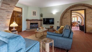 A living room with a fireplace, a television, and a dining area visible through an archway. - Podere Gellino 14, Emma Villas (Chianciano Terme)
