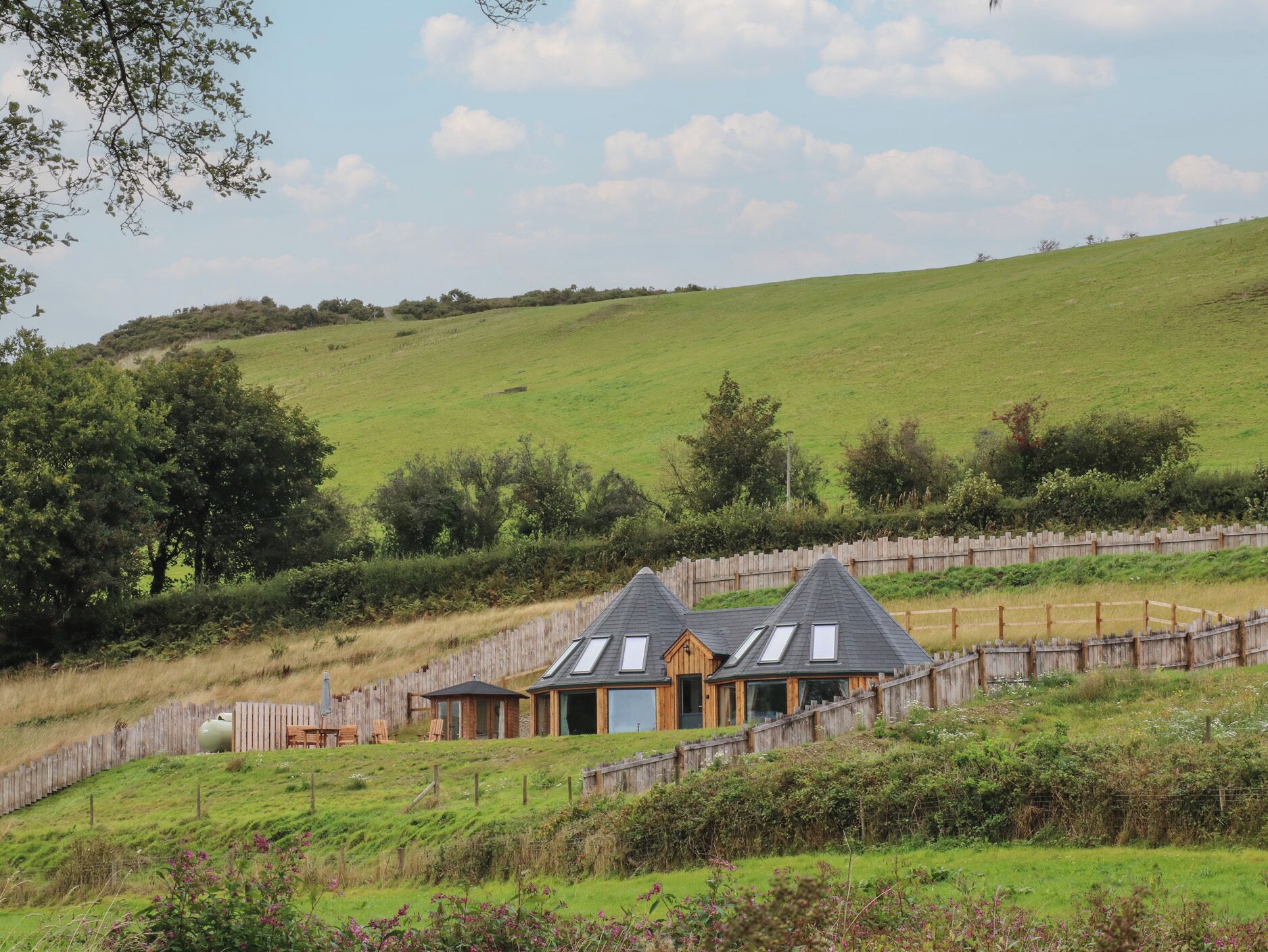 A rural setting with two wooden houses on a hillside, surrounded by a wooden fence and lush greenery.