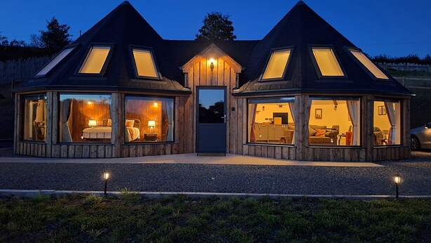 Une cabane moderne, dotée de plusieurs pièces, de grandes fenêtres et d’une porte centrale, entourée d’une allée en gravier et d’un espace paysager aménagé.