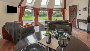 Un coin repas avec une table ronde, une bouteille de vin et un verre de vin rouge. Il y a deux fenêtres avec des rideaux, un téléviseur accroché au mur et une vue sur un paysage verdoyant.