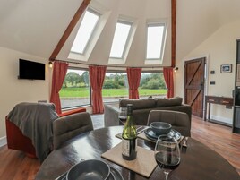 A dining area with a round table, a bottle of wine, and a glass of red wine. There are two windows with curtains, a television on the wall, and a view of a green landscape.