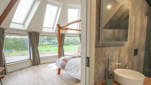 A bedroom with a large skylight, a wooden bed, a bedside table, and a bathroom with a sink and mirror. - Yr Awen - Inspiration (Lampeter)