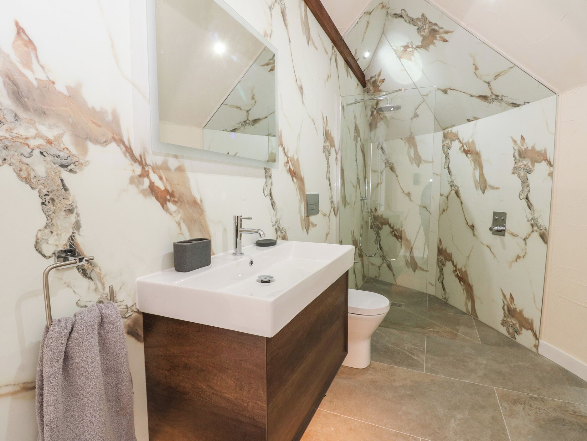 A modern bathroom with a marble wall, a large mirror, a wooden vanity, a shower enclosure, and a toilet.