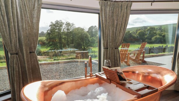 Tinaja de cobre llena de hielo, ubicada junto a una ventana grande con cortinas, con vistas a un espacio al aire libre cubierto de césped, con sillas y una mesa de madera.