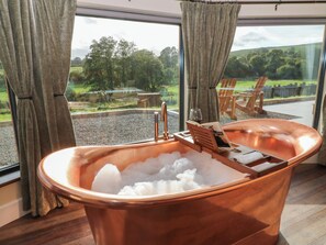 A copper hot tub filled with ice, positioned by a large window with curtains, overlooking a grassy outdoor area with wooden chairs and a table. - Ty Crwn - The Roundhouse (Lampeter)