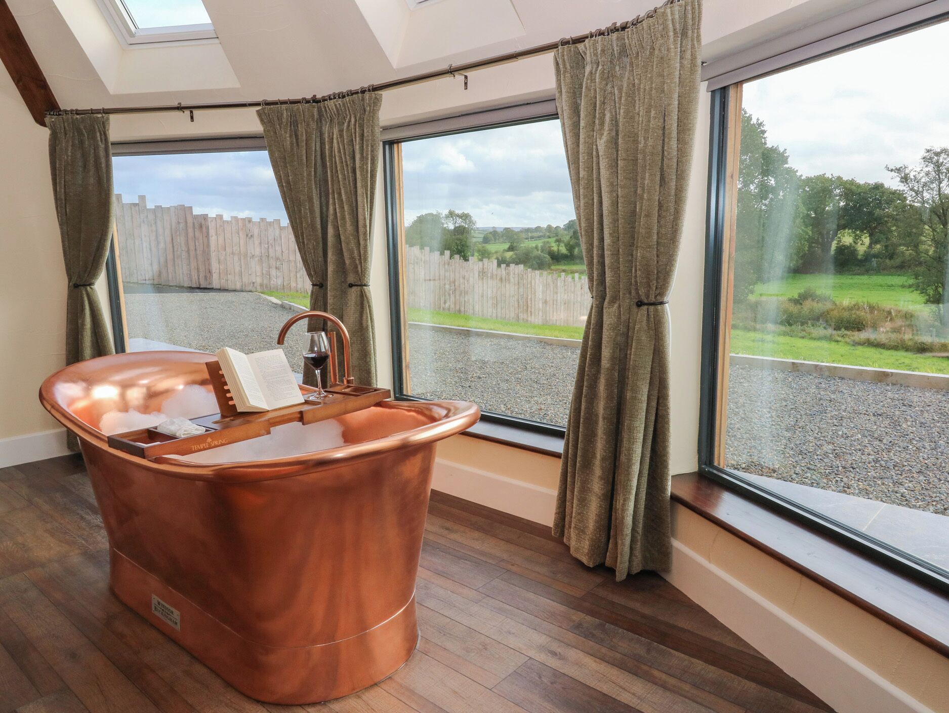 Un baño moderno con una bañera de cobre, ventanas amplias y vistas a una valla de madera y follaje.