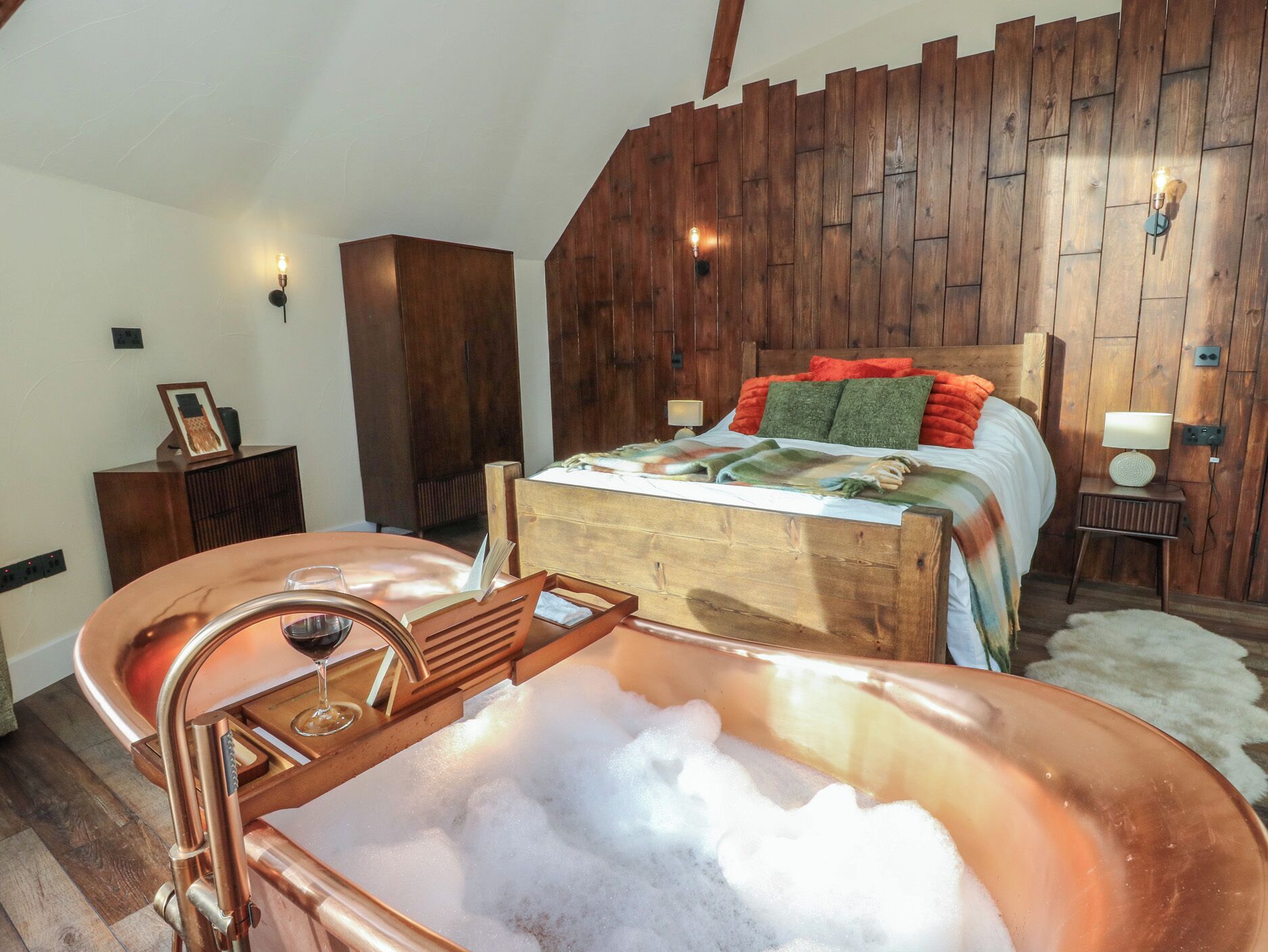 A bedroom with a wooden headboard, a large bathtub filled with bubbles, a wooden nightstand with a lamp, and a wooden wardrobe.