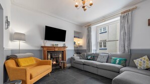 A living room with a fireplace, a sofa, a TV, and a window with curtains. - Lan Y Mor - Close to Town and Beach (Tenby)
