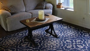Ein gemütliches Zimmer mit Holzbalkendecke, einem grauen Sofa, einem kleinen Tisch mit Kerze und einem gemusterten Teppich.