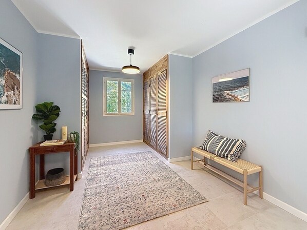 Een hal met een lichtblauwe wand, een houten bankje, een kamerkleed met patroon, een kleine tafel met een plant en een raam met luiken.