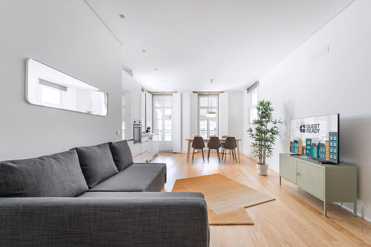 Ein modernes Wohnzimmer mit grauem Sofa, einem Essbereich mit Tisch und Stühlen, einem Fernseher auf einer Kommode und einem großen Spiegel an der Wand.