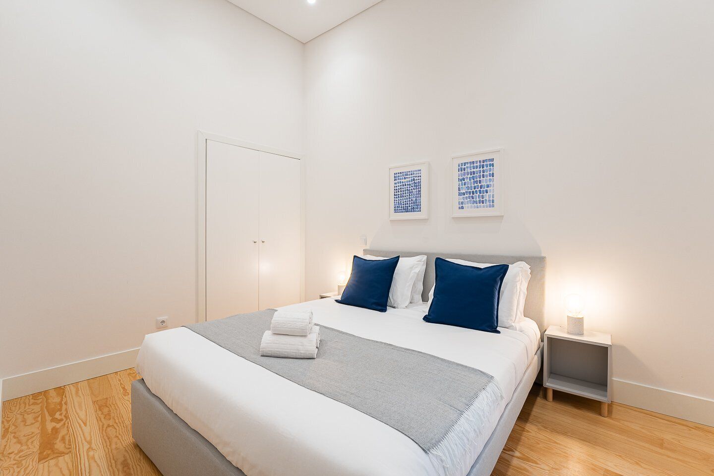 A modern hotel room with a large bed, a bedside table with a lamp, and two wall art pieces.