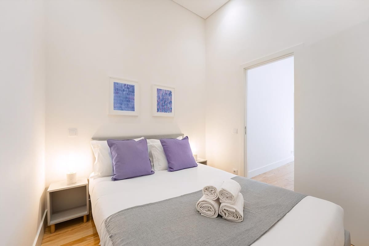Ein ordentlich bezogenes Bett mit grauer Decke und zusammengerollten Handtüchern, ein Nachttisch mit Lampe sowie zwei gerahmte Kunstwerke an der Wand.