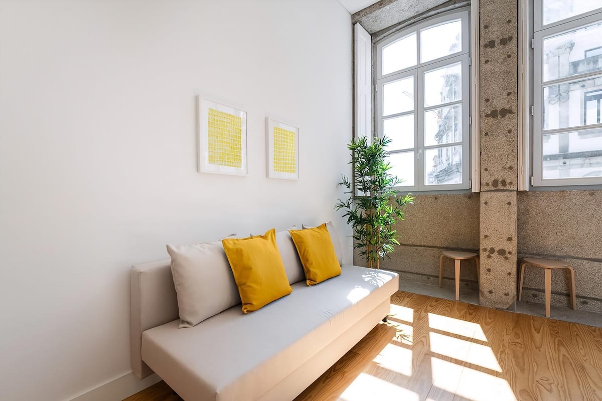 Ein modernes Wohnzimmer mit einer Couch, zwei gelben Kissen, einer Topfpflanze und einem Fenster mit Blick auf Gebäude.