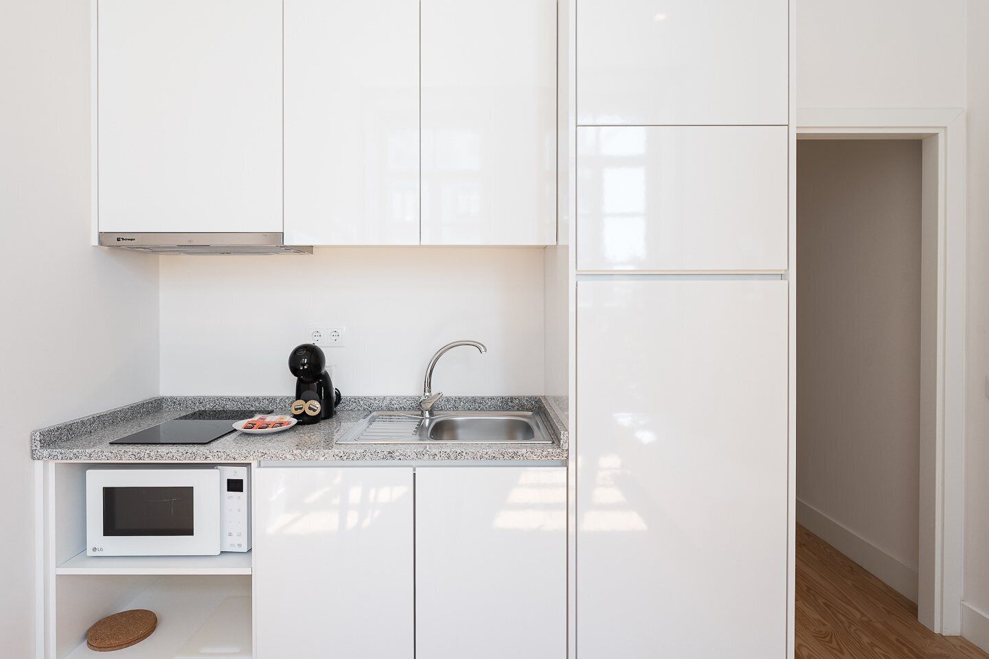 A compact kitchen with white cabinets, a granite countertop, a microwave, a sink, and a black coffee machine.