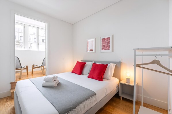 Una camera d'albergo moderna con un letto grande, un comodino con una lampada, una sedia e vista sulla città attraverso un'ampia finestra.