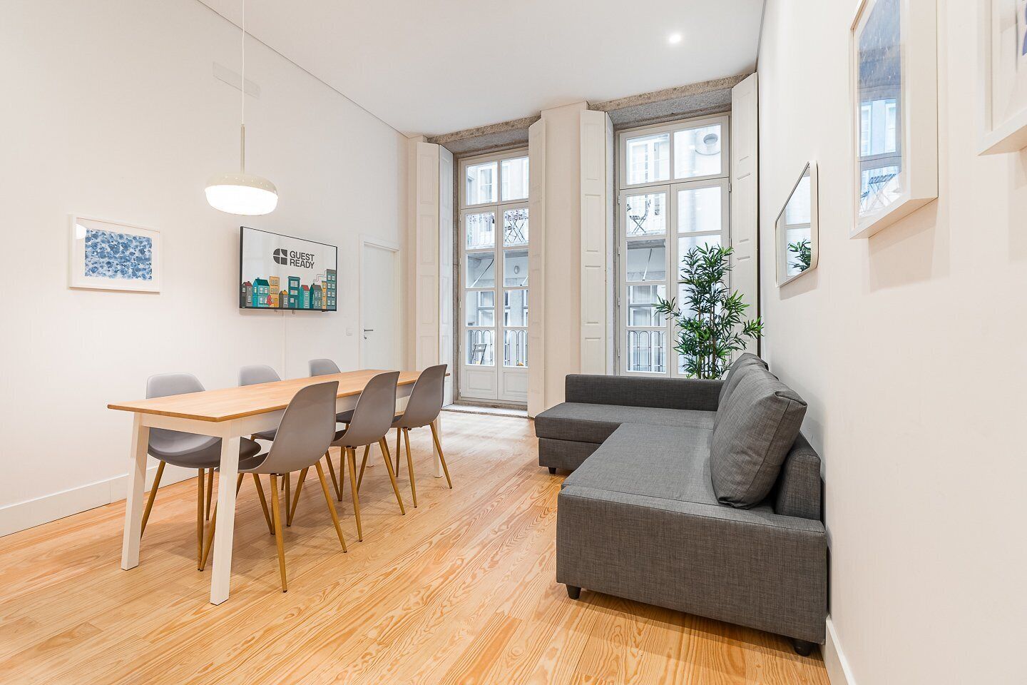 A modern living room with a grey sofa, wooden dining table, and chairs. There are large windows and a plant in the corner.