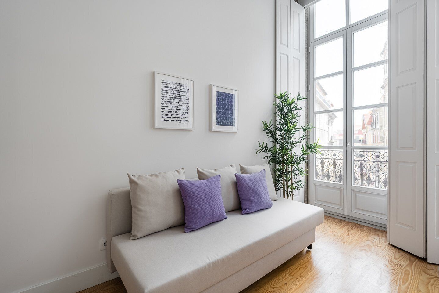 Ein modernes Wohnzimmer mit einem beigefarbenen Sofa, zwei gerahmten Kunstwerken und einem großen Fenster mit Blick auf Gebäude.