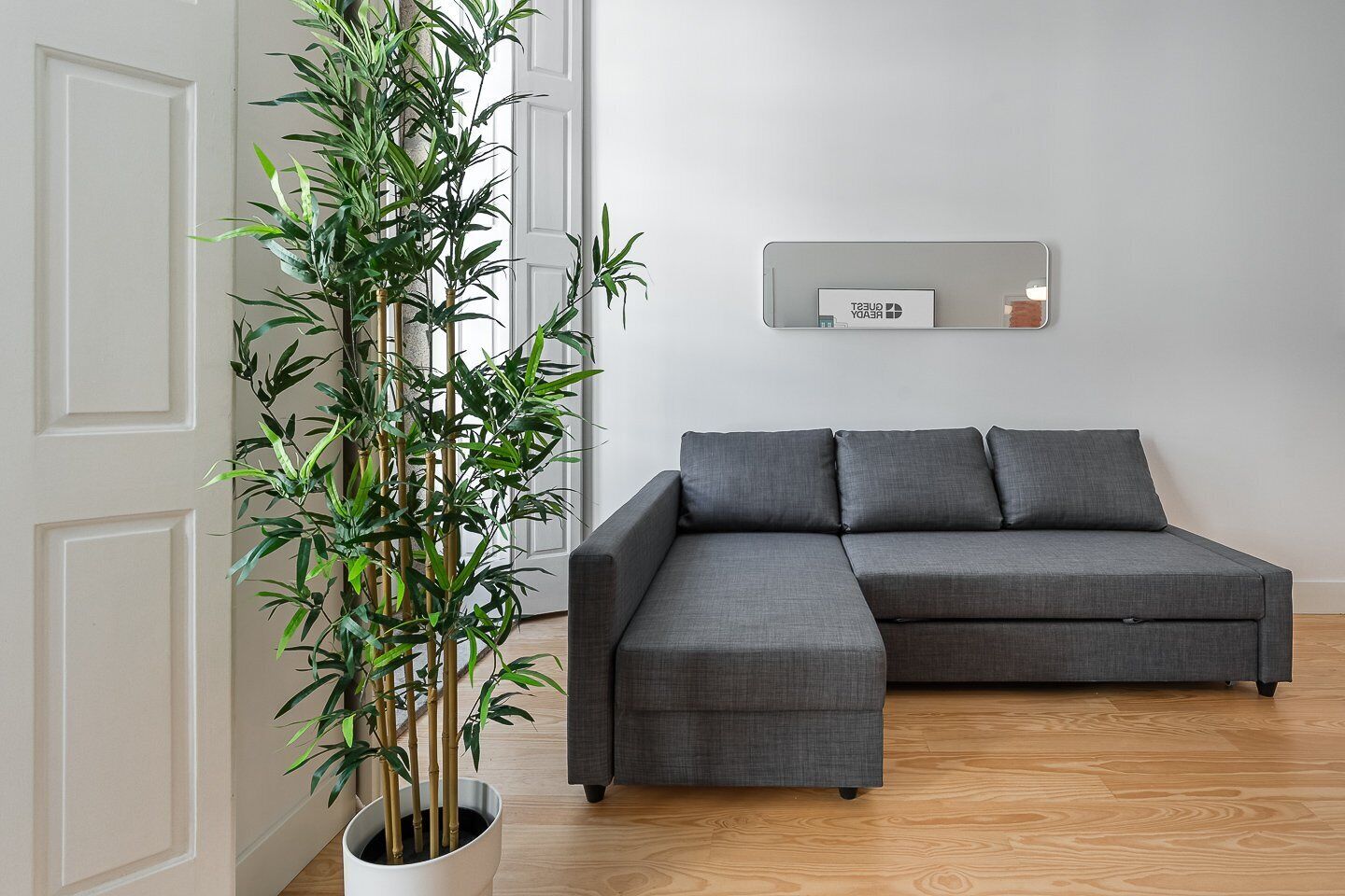 A modern living room with a grey sectional sofa, a tall bamboo plant in a white pot, and a wall-mounted mirror with a 'REUSABLE' sign.