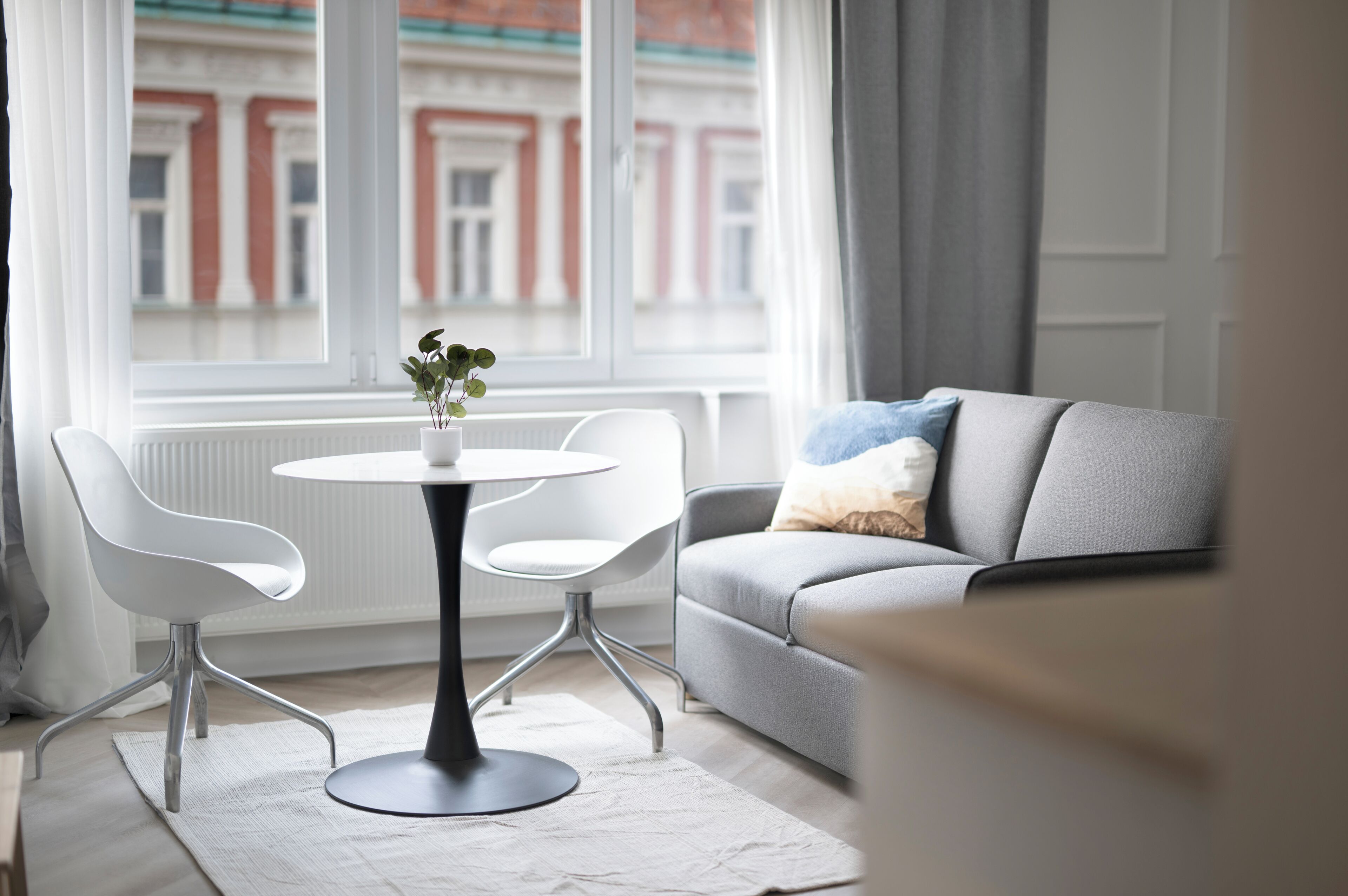 A modern living room with a grey sofa, white chairs, a round table with a vase, and a view of a building through a window.
