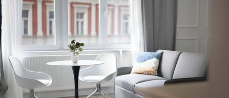 Una sala moderna con un sofá gris, sillas blancas, una mesa redonda con un florero y vistas a un edificio a través de una ventana.