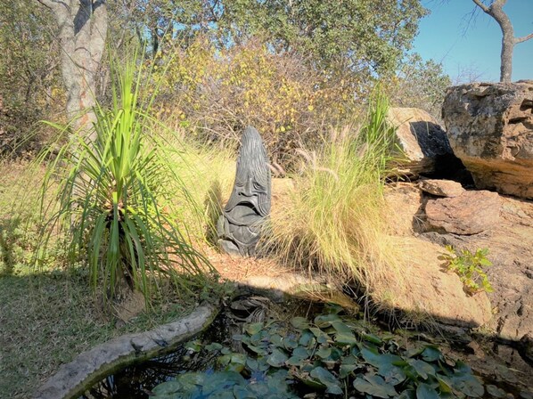 A natural outdoor setting with a rock formation, a carved wooden figure, and surrounding vegetation. - Double Lodge in Natural African Bush - 2111 (Bulawayo)