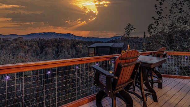Puinen terassi, jossa on pöytä ja tuoleja, ja josta avautuu näkymä vuoristomaisemaan auringonlaskun aikaan.