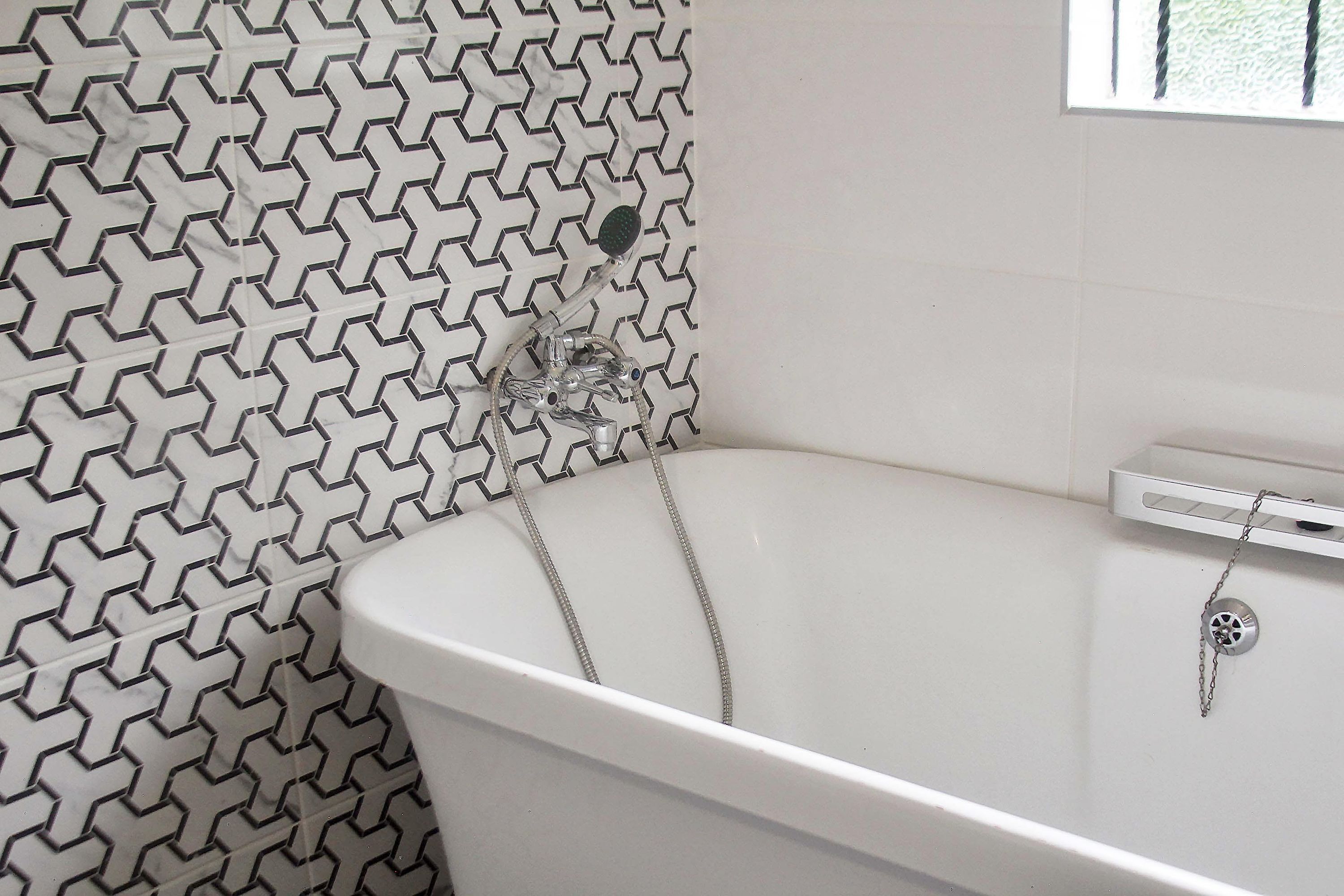 Una bañera blanca con ducha y panel de control, frente a una pared con diseño geométrico.