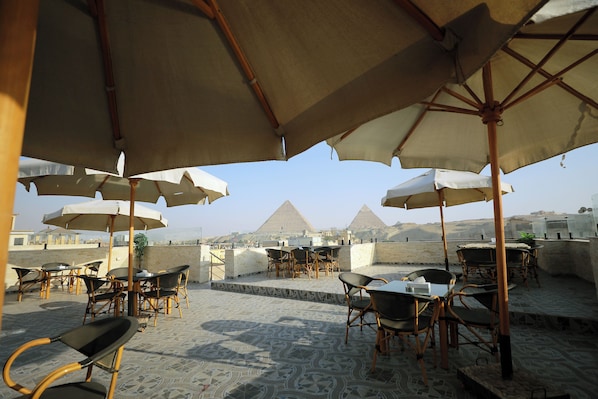 An outdoor seating area with tables and chairs under umbrellas, overlooking a pyramid.